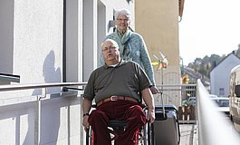 Mann im Rollstuhl auf einer Rampe im Eingansbereich eines Gästehauses, dahinter eine ältere Frau, die einen Koffer hinter sich herzieht